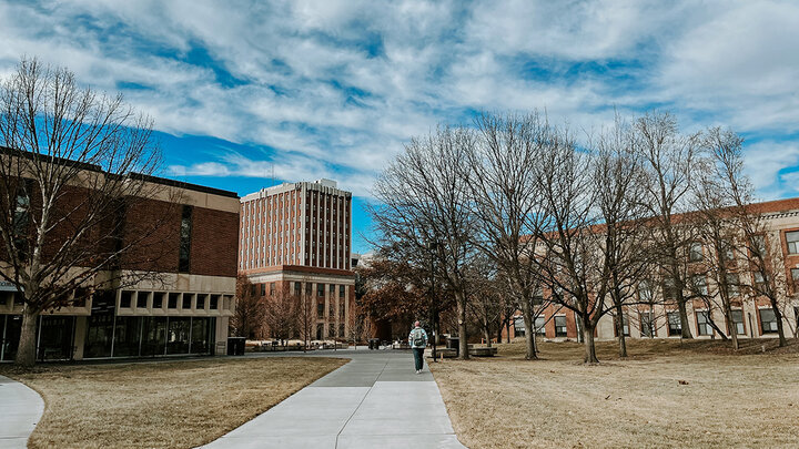 A student walks down a concrete path with three red brick buildings in the background.